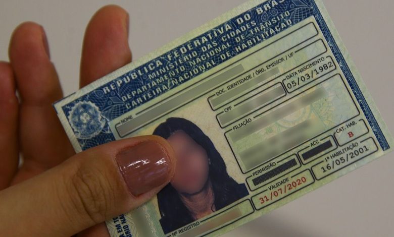 Carteira Nacional de Habilitação (CNH) brasileira sendo segurada por uma pessoa, com informações parcialmente borradas.
