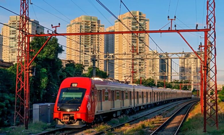 Trem da Linha 12-Safira circulando em área urbana com edifícios ao fundo e rede elétrica aérea.