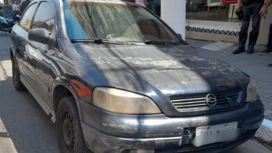 Astra azul em mau estado, apreendido por monitoramento inteligente de trânsito, com policial ao fundo em São Paulo.