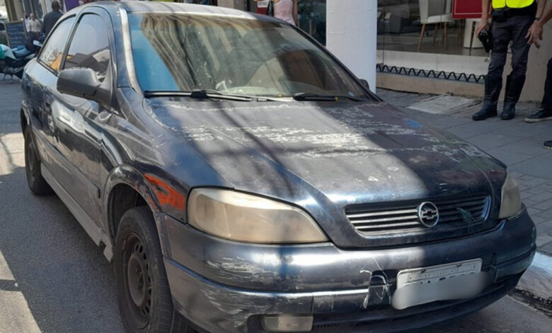 Astra azul em mau estado, apreendido por monitoramento inteligente de trânsito, com policial ao fundo em São Paulo.