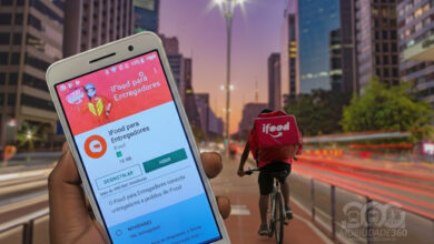 Mão segura celular com app do iFood em frente a um entregador ciclista, representando a rotina que usará o chip iFood para entregadores.