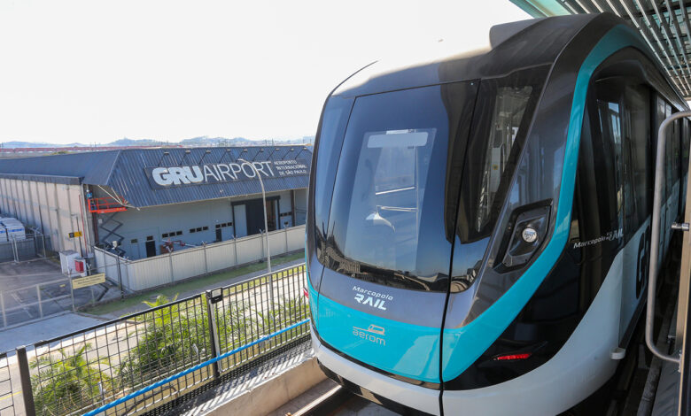 Aeromóvel de Guarulhos estacionado em trilhos elevados, com o edifício GRU Airport ao fundo. O projeto está paralisado.