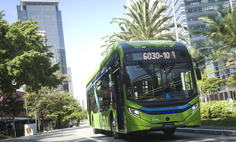 Ônibus elétrico Mercedes-Benz eO500 U verde circulando em avenida urbana de São Paulo, com prédios ao fundo.